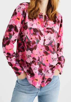 Vrouw draagt een langärmelig overhemd met knopen met een felroze, paarse en rode bloemenpatroon en lichtblauwe spijkerbroek, handen in de zakken.