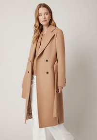 Kamelowy płaszcz dwurzędowy wykonany z mieszanki wełny, z wyprofilowanym kołnierzem, bocznymi kieszeniami i guzikowymi detalami, w połączeniu z kremowym topem i spodniami.