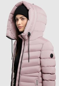 Kvinde iført lyserød polstret hættejakke og sort strikket hue, der vender til venstre, på en ensfarvet lys grå baggrund.