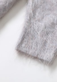 Dettaglio di un maglione lavorato a maglia grigio chiaro con una consistenza morbida e pelosa. Presenta polsini a costine e sottili variazioni di colore nel filato.