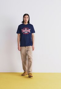 Marineblå bomull T-skjorte med rød "Reebok Classic" grafikk, kombinert med beige bukser og brune sko, plassert mot en gul bakgrunn.