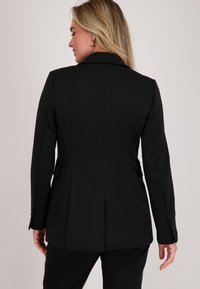 Vrouw met blond haar die een zwarte, op maat gemaakte blazer en bijpassende broek draagt, gezien van achteren tegen een eenvoudige neutrale achtergrond.
