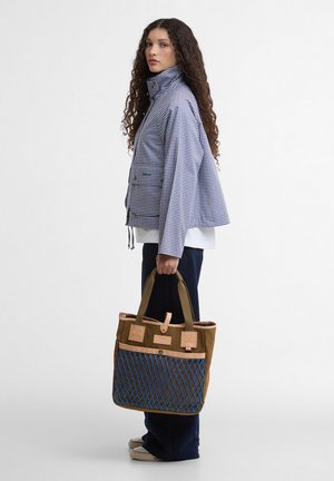 Γυναίκα με μακριά, σγουρά μαλλιά, που φοράει ένα μπλε καρό σακάκι και σκούρο παντελόνι, κρατώντας μια καφέ τσάντα ώμου με μπλε δίχτυ.