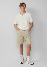 Lichtgewicht wit gestreept poloshirt met een V-hals, gecombineerd met beige shorts en beige sneakers. Model staat met de handen in de zakken.