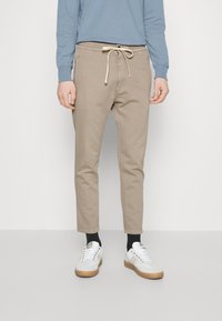 Beige, taps toelopende broek met een elastische tailleband en trekkoord, voorzien van een effen kleur en subtiele textuur. Gecombineerd met witte sneakers en zwarte sokken.