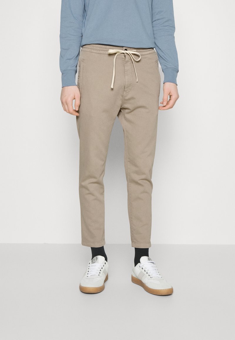 Beige, taps toelopende broek met een elastische tailleband en trekkoord, voorzien van een effen kleur en subtiele textuur. Gecombineerd met witte sneakers en zwarte sokken.