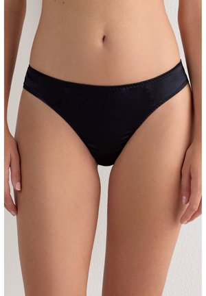 Kvinde iført sorte satin bikini-trusser med mellemhøjt liv, stående med hænderne afslappet ned langs siderne mod en ensfarvet baggrund.