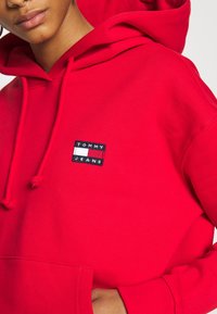 Czerwona bluza z kapturem wykonana z bawełny, z przednią kieszenią typu kangur i czarną metką z haftowanym logo "TOMMY JEANS" na klatce piersiowej.