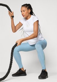 Hvitt kortermet treningsskjorte, lyseblå leggings med grønn tekst, og svarte joggesko. Modellen holder et tykt, svart treningsrep.