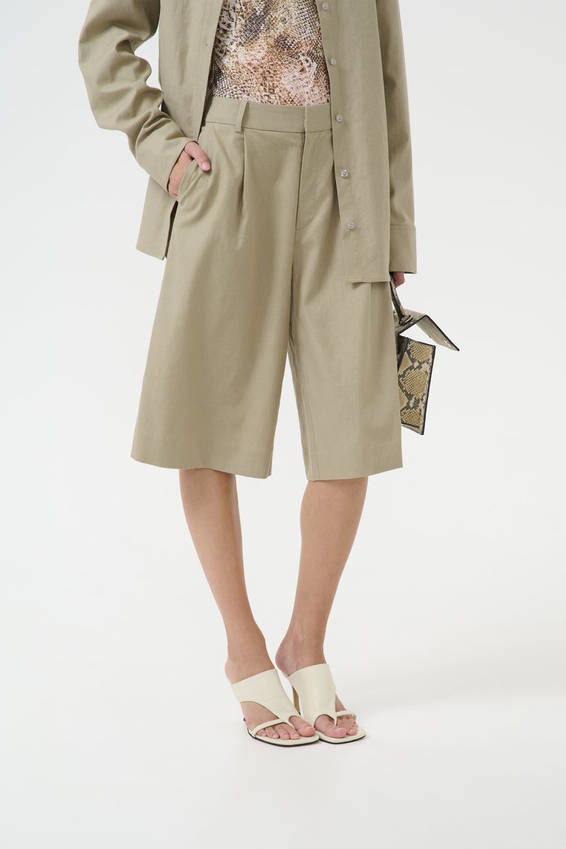 Shorts beige ajustés avec des plis, associés à un haut à motifs et des sandales blanches à enfiler. Les accessoires comprennent un petit sac texturé.