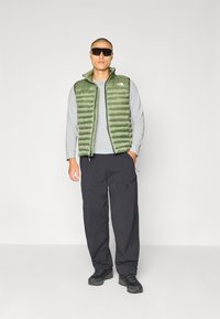 Groene puffer vest met een geruite stijl over een grijze langarmshirt, gecombineerd met zwarte cargo broek en zwarte wandelschoenen.