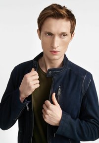 Granatowa skórzana kurtka z perforacjami, z zamkiem z przodu, zatrzaskowym kołnierzem i teksturowanymi akcentami. Nosi klasyczny oliwkowy podkoszulek.