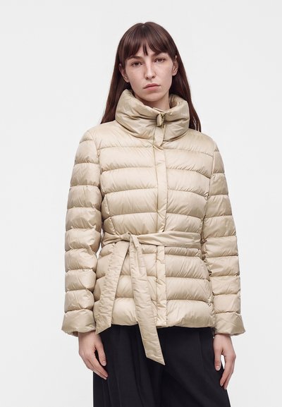 Frau mit glatten braunen Haaren, die eine beige Steppjacke mit hohem Kragen und gebundenem Gürtel trägt, vor einem schlichten weißen Hintergrund stehend.