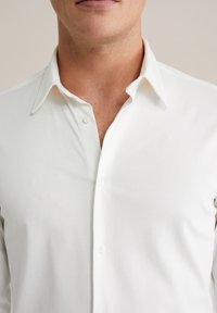 Uomo che indossa una camicia bianca col colletto sbottonato, mostrando parte del collo e della parte inferiore del viso con una leggera barba.