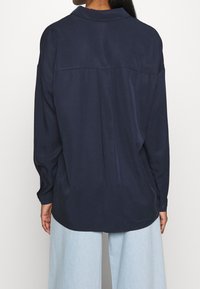 Námořnická modrá košile s dlouhým rukávem a límečkem, volnější střih, se středovým švem na zadní straně. Tkanina vypadá hladce s mírným leskem.