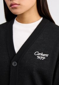 Cardigan preto feito de tecido de malha, com decote em V, fecho de botão único e o logotipo bordado em branco "Carhartt WIP" no peito.
