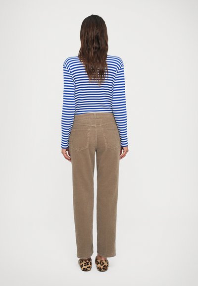 Vrouw die met de rug naar de camera staat, gekleed in een blauw-wit gestreepte longsleeve top, bruine corduroybroek en schoenen met luipaardprint.