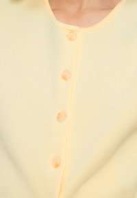 Ανοιχτό κίτρινο ζακέτα με λαιμόκοψη V, ριμπ υφή και έξι κουμπιά στο ίδιο χρώμα κατά μήκος του μπροστινού μέρους για κλείσιμο.