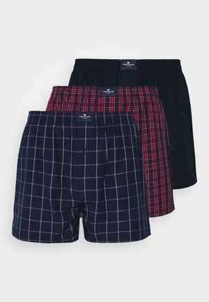 Trois paires de boxers empilées : bleu marine avec un motif grille blanc, écossais rouge, et noir uni. Chacun a une taille élastique et un logo.