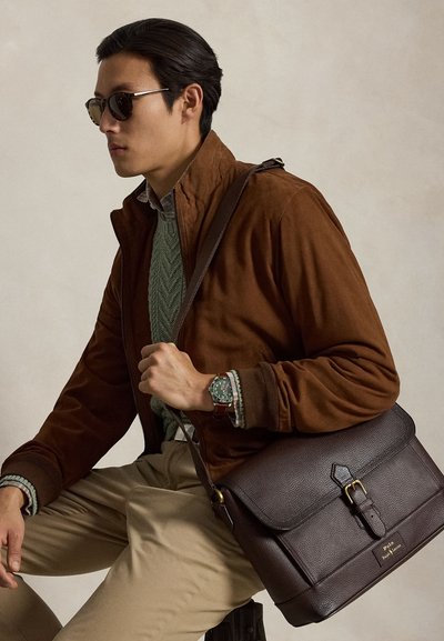 Άνδρας με γυαλιά ηλίου, καφέ μπουφάν, πράσινο πουλόβερ, μπεζ παντελόνι, κρατώντας σκούρα καφέ δερμάτινη τσάντα ώμου και φορά ρολόι χειρός.