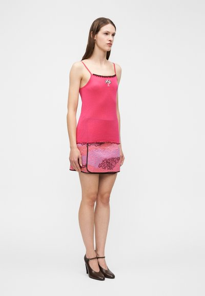 Γυναίκα που φοράει ροζ καλοκαιρινή μπλούζα με μικρό σχέδιο δέντρου και μίνι φούστα με μοτίβο, συνδυασμένα με σκουρόχρωμα παπούτσια στιλέτο, όρθια πλάγια.