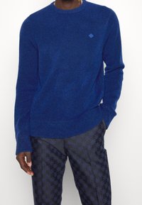 Blå stickad tröja med rund hals, strukturerad tyg och en liten diamantlogotyp på bröstet. Bärs med mönstrade mörka byxor.