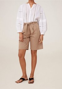 Valkoinen pitkähihainen pusero, jossa on leikkauskoristeita, yhdistetty korkeavyötäröisiin beigeihin shortseihin, jotka on sidottu vyötäröltä. Päällä mustat varvassandaalit.