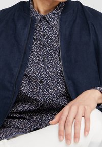 Námořnická bombarďácká bunda s hladkou texturou, vybavená zipem a vroubkovaným límcem, vrstvená přes námořnickou košili s drobným květinovým vzorem.