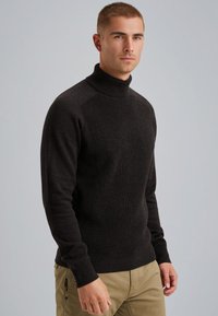 Un pull noir encolure haute en tricot texturé, avec des manches longues et une silhouette ajustée. Porté avec un pantalon khaki.