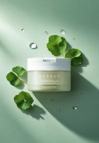 Läpinäkyvä astia valkoisella kannella, sisällä vihreä tuote. Ympäröity vihreillä lehdillä ja vesipisaroilla vaaleanvihreällä taustalla.