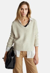 Béžový žebrovaný svetr s v ýstřihem do V, vrstvený přes černý top a bílou pruhovanou košili, v kombinaci se světle hnědými širokými kalhotami a černou kabelkou.