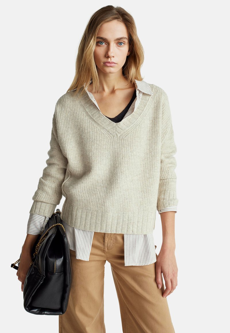 Béžový žebrovaný svetr s výstřihem do V, vrstvený přes černý top a bílou pruhovanou košili, v kombinaci se světle hnědými širokými kalhotami a černou kabelkou.