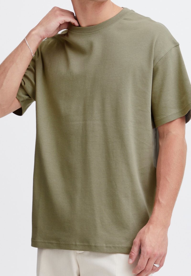 Homme portant un t-shirt vert olive à manches courtes avec un pantalon beige, touchant le col avec la main gauche, montrant un bracelet et une bague en argent.