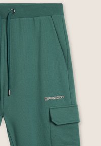 Pantalones de chándal verdes hechos de tela suave con cintura con cordón, bolsillos laterales y un bolsillo tipo cargo que presenta un detalle del logo plateado "FREDDY".