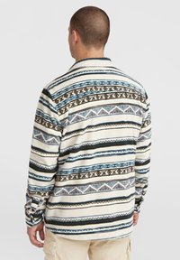 Sweter polarowy z długimi rękawami, w kolorze kremowym, z niebieskimi, brązowymi i czarnymi geometrycznymi wzorami. Posiada luźny krój i mankiety zapinane na guziki.