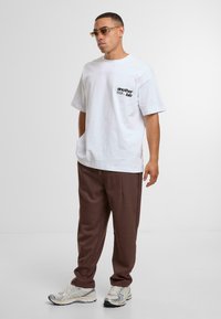 Tricou alb din bumbac cu un logo grafic negru pe pieptul stâng, asortat cu pantaloni maro evazați și teniși argintii.