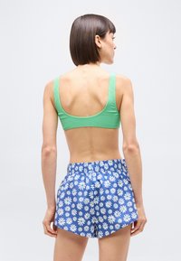 Γυναίκα π ου κοιτάζει προς τα πίσω, φοράει πράσινο crop top και μπλε σορτς με λευκά λουλούδια μαργαρίτας, στέκεται μπροστά σε λευκό φόντο.