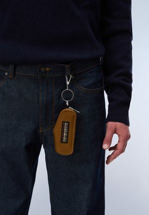 Person trägt dunkelblaue Jeans und schwarzen Pullover mit brauner Napapijri-Schlüsselmappe, die an der Gürtelschlaufe auf der rechten Seite befestigt ist.