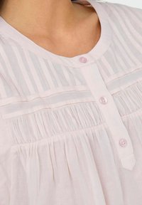 Jasnoróżowa bluzka z plisowanym przodem, okrągłym kołnierzykiem i różowymi guzikami. Materiał jest miękki i lekki z wodzącymi detalami.