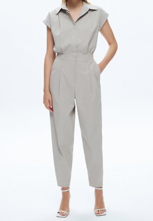 Combinaison gris clair à manches courtes, col à revers et jambes fuselées. Présente des rayures verticales discrètes et des poches latérales. Sandales visibles.