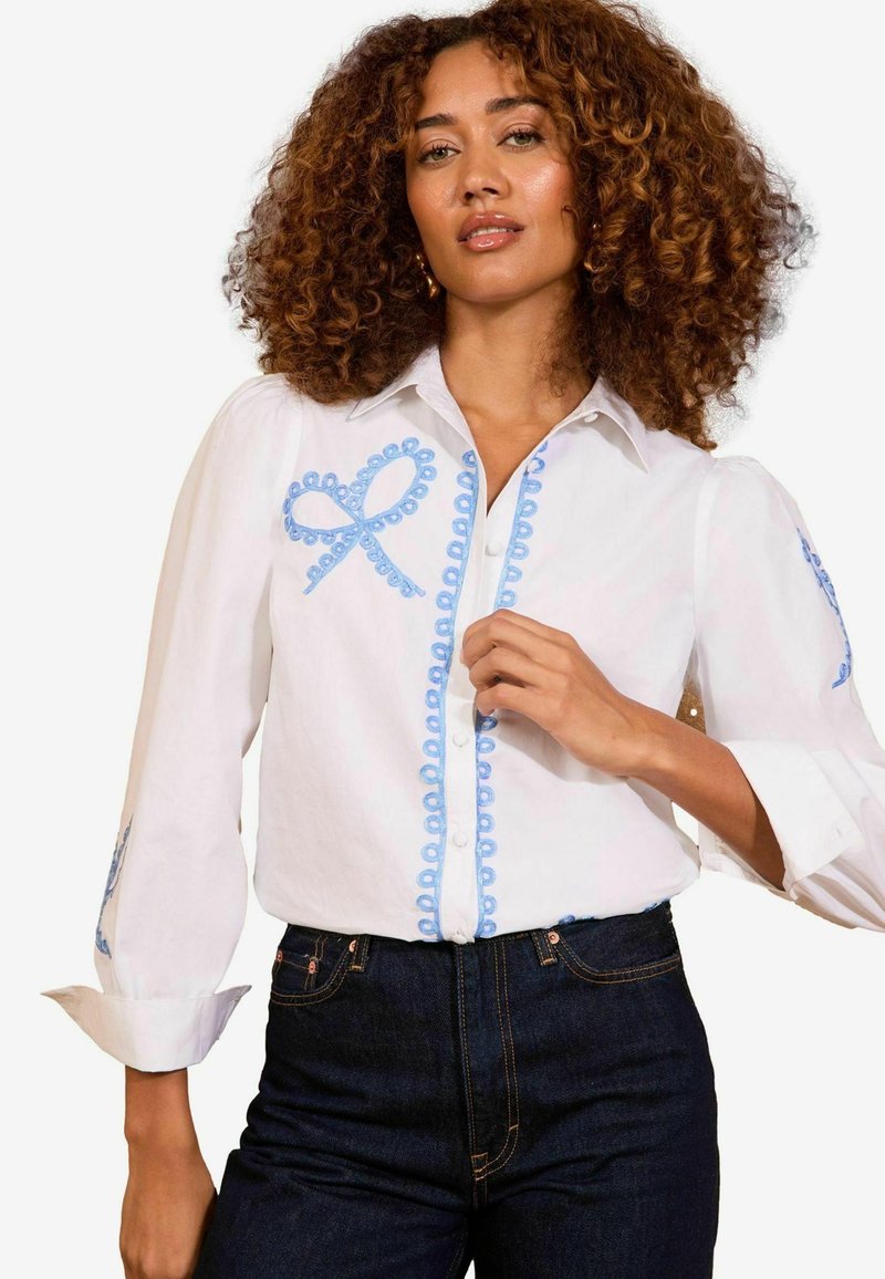 Camicia bianca con bottoni, caratterizzata da ricami blu a forma di anelli e dettagli, colletto classico e maniche lunghe leggermente gonfiate. Jeans in denim visibili.