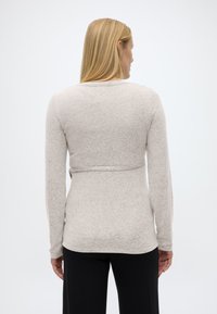 Γυναίκα με ίσια ξανθά μαλλιά φοράει ανοιχτό γκρι μπλουζάκι μακρυμάνικο και μαύρο παντελόνι, γυρισμένη προς τα πίσω μπροστά σε λευκό απλό φόντο.