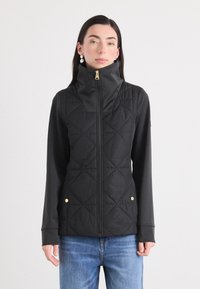 Een jonge vrouw poseert in een zwart gewatteerd vest met een hoge kraag, gecombineerd met lichtblauwe jeans, tegen een effen witte achtergrond.