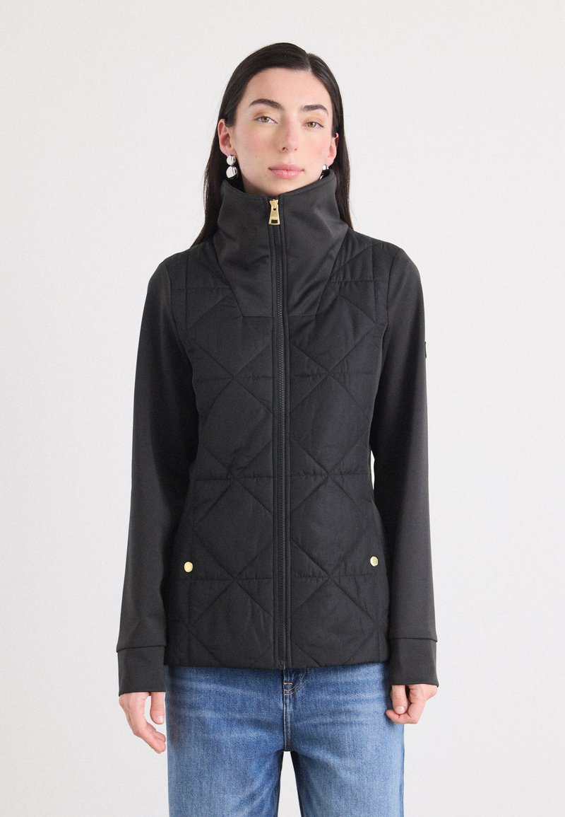 Een jonge vrouw poseert in een zwart gewatteerd vest met een hoge kraag, gecombineerd met lichtblauwe jeans, tegen een effen witte achtergrond.