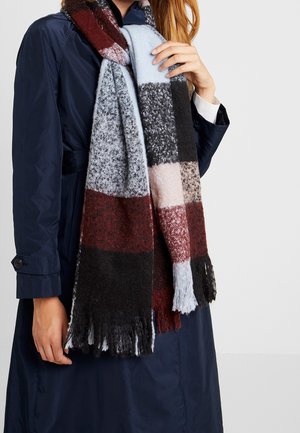 Personne portant un manteau bleu marine et une écharpe à carreaux multicolores avec des blocs noirs, bordeaux, gris et bleu clair, à extrémités frangées.