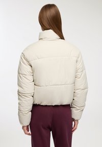 Nadmuchiwana beżowa kurtka z wysokim kołnierzem, z poziomym przeszyciem, krótszym fasonem i gładką fakturą, w połączeniu z bordowymi spodniami.