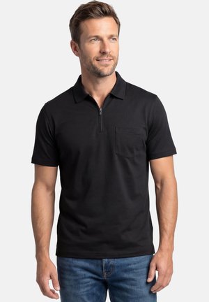 Uomo che indossa una polo nera a maniche corte con tasca sul petto e cerniera, abbinata a jeans blu, che guarda leggermente verso destra.