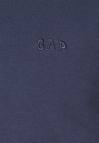 Tumši zilā kokvilnas auduma ar strukturētu apdari. Uz auduma centrā atrodas izšūts "GAP" logo nedaudz tumšākā tonī.