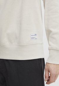 Sudadera color crema con puños y dobladillo acanalados, que presenta una etiqueta de tela en la esquina inferior izquierda con texto en negro. Pantalones oscuros parcialmente visibles.