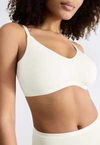 Donna con capelli ricci che indossa un crop top bianco senza cuciture e pantaloni a vita alta coordinati, su uno sfondo leggero e neutro.